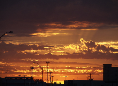 [A closer view of the city buildings a bit later in the morning shows rays of the sun hits the underside of the big cloud. The sun is still below the horizon and the skay is a profusion of yellow and orange.]